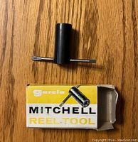 Fishing reel tool shown with original yellow and white Mitchell box on wooden surface.