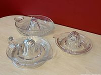 Three clear glass manual juice reamers on light wooden surface against red background, showing bowl shape and handles