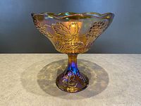 Front view of marigold carnival glass footed fruit bowl showing embossed grape and leaf details and scalloped rim.
