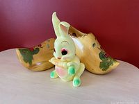 Plastic vintage bunny toy sitting upright, cream-colored with pastel green feet and ears, pink belly, and large painted eyes, featuring a ruffled collar detail around its neck.