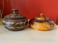 Two vintage carved lidded trinket dishes shown side by side on table in front of red wall. One dark wood with carved leaves, one light brown ceramic with carved leaves and shapes.