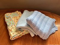 Top view of three folded children's blankets on a wooden surface, showing the crochet blanket, textured blanket, and quilted blanket with animal prints.