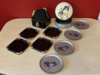 Photo showing two vintage drink coaster sets: six black square coasters with gold corners and a black holder, plus five round silver coasters with blue flower design and a silver holder stand