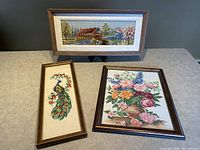 Overall view of three framed needlepoint artworks: covered bridge, peacock, and floral bouquet.