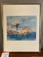 Framed watercolour artwork showing a coastal scene with blue sea and sky, earth tone buildings, in a simple gold-tone metal frame with matting.