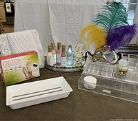 Overview photo showing bath pillow, detox foot pads box, makeup storage unit, Mardi Gras mask, assorted bottles of wine-scented bath salts on tray, white drawer storage unit, moisturizer jar, and glass bottle with USB light.