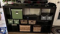 Front view of black wooden open shelved cabinet holding green IKEA boxes, black and white striped boxes, and basket storage