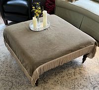 Full view of square ottoman covered with brown velvet-like fringed fabric, showing wooden legs and placement of tray and candles on top.