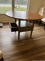 Full view of round drop leaf table with drawer, wood top and greenish distressed base.
