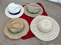 Photo showing all four hats arranged on a red cloth: white fedora, beige woven fedora, knit lacy sunhat, natural woven floppy hat with turquoise band.