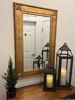 Mirror with golden wooden frame, two lanterns with candles, and small faux pine tree on wooden surface.