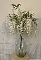 Full view of the large clear glass vase filled with white silk wisteria flowers and green leaves, showing the overall height and shape of the lot.
