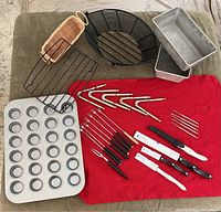Photo showing all kitchen items including CUTCO knives, lobster crackers, fondue forks, muffin tin, loaf pans, roaster rack, wire basket, and bamboo bread holder on a red cloth and green surface.