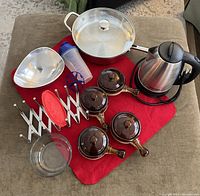 All items arranged on a red cloth showing ceramic bowls with lids, electric kettle, large metal strainer, Pyrex bowl, lid holder, and frying pan.