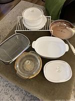 Photo showing all kitchen items laid out including Copper Chef frying pan lid, Corning Ware dishes with lids, Anchor casserole and basket, wire cabinet dish rack, and sink strainer.