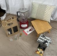 Full lot view showing wooden stool, KitchenAid blender, wine carrier bag, shot glasses, pasta maker, and Pier 1 pillow.