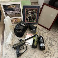 Overview of paintball gear and framed sports memorabilia arranged on floor including paintball gun, mask, loader, two compressed air tanks, two framed prints, empty frame.