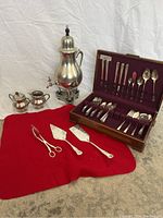 Overview photo of the complete lot showing the pewter tea set including tea urn, creamer, sugar bowl, the red cloth, assorted silver plated utensils, and the cutlery box open with cutlery inside.