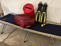 Full lot showing LLBean easy cot, red sleeping bag in sack, maroon luggage, black and yellow fins, unopened tennis ball can