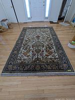 Full view of the rectangular area rug laid out on a hardwood floor, showing the delicate floral and animal motifs, fringe edges, and overall condition.