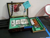 Photo showing wooden jewelry box with mirror, containing necklace, pins, and watches arranged inside compartments; also shows an adjacent tray with orange and black beaded necklace and other pins.