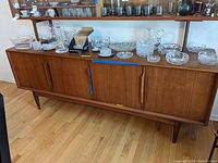 Wide view of shelf with multiple cut glass and crystal items including pitchers, bowls, platters, decanter, dishes, ice bucket and coasters displayed on top of a wooden cabinet.