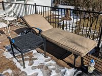 Overall view of cast aluminum chaise lounge with beige cushion and matching side table on outdoor deck with snow patches