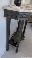 Side angle showing marble top, carved wood apron with Greek key design, tapered legs, and lower shelf with decorative item.
