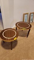 Two round mid-century style side tables with laminate tops and tile inlay rings, standing on carpeted floor next to framed artwork.