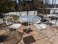 Full patio set on outdoor deck showing glass-top table with four matching chairs in white metal with white cushions.