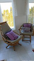 Two rattan chairs (scoop and club) positioned by windows, each with gray cushions and purple decorative pillows.