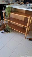 Full view of a three-tier wooden shelf with cylindrical legs and two plants placed on it, located in a tiled room.