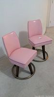 Two pink vinyl padded swivel chairs with padded seats and backs, metal circular footrest, and brown legs shown from the front and angled sides.