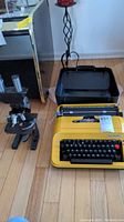 Photo showing the yellow Privileg 350 T typewriter next to the Bausch & Lomb microscope on a wooden floor, with black carrying case open behind the typewriter.