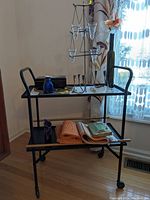 Full view of vintage black and brass metal rolling cart with two shelves holding assorted items including candle holders, cobalt bottle, keepsake box, candles, trivets, and woven mats.