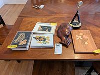 Photo showing the wooden floral marquetry panels, framed needlepoint butterfly artworks, carved wooden wall bust, and black Seiko clock on a wooden table.