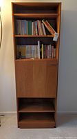 Front view of tall wooden cabinet/bookcase with three open shelves holding books and closed lower section with door.
