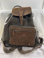 Front view of the dark brown leather backpack showing the suede-textured flap with buckle closure and front zipper pocket.