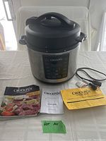 Photo showing Crock-Pot Express multi-cooker along with three manuals: recipe book, owner's guide, and additional manual, plus power cord on a table.