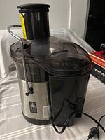 Side and angled view of the Jack LaLanne juicer displaying its stainless steel body, plastic top with locking arm, attached juice spout, and power cord.