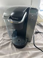 Front and side view showing the Keurig logo, digital screen on right panel, large transparent water reservoir on left, and power cord.