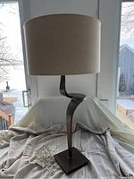 Full view of the bronze base table lamp with beige linen-look shade on a fabric surface by a window.