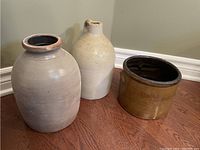 Image showing three stoneware crocks: two jug-shaped and one round, positioned on wooden floor and against wall.