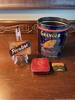 Photo showing the four advertising tins including the large Granger tobacco tin and the smaller Picobac, Cotton Coupon Paper Co., and Canada Motor Products Ltd. tins.