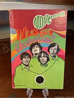 Front cover of The Monkees "Who's Got The Button" hardcover book featuring colorful psychedelic design and an illustration of the four band members.