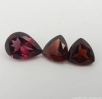 Three garnet gemstones displayed on a plain white background, showing a pear-shaped garnet and two smaller stones including a trillion cut with faceting visible.