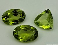 Three peridot gemstones on a white background, showing two oval-shaped and one triangular-shaped stones with faceted cuts.