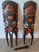 Full front view of pair of South African carved wooden tribal masks with complex heart-shaped and geometric painted details. One mask shows a separate carved piece lying in front.