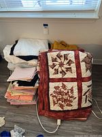 Photo showing variety of bedding items including a red and cream patterned quilt, assorted folded sheets, and electric hot pads with cords