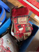 Blue plastic bin containing a red and green fabric holiday stocking with a snowman and 'Let It Snow' text, among other decorations.